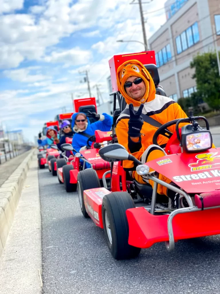 IMG_2813 - StreetKart Person in an orange mascot hoodie and sunglasses drives a red go-kart at the front, making a peace sign as a line of riders in colorful outfits follows on a city street.”,