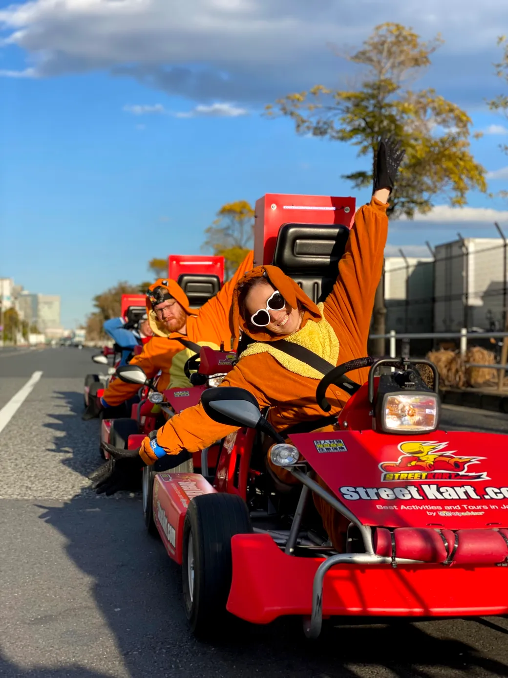 Person in an orange costume waves from a bright red go-kart during a street parade, wearing white sunglasses and a black glove on the raised arm.