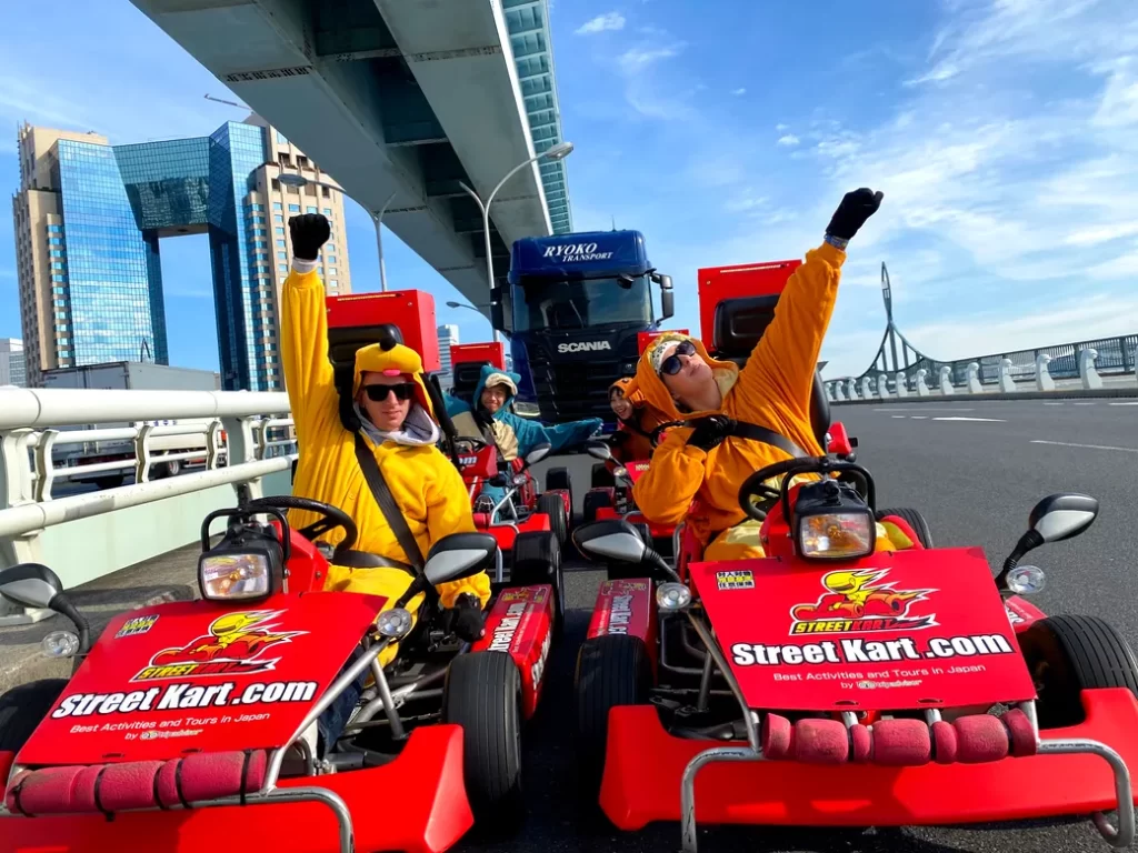 Group of people in bright yellow and orange outfits riding red go-karts on a city bridge, waving at the camera.